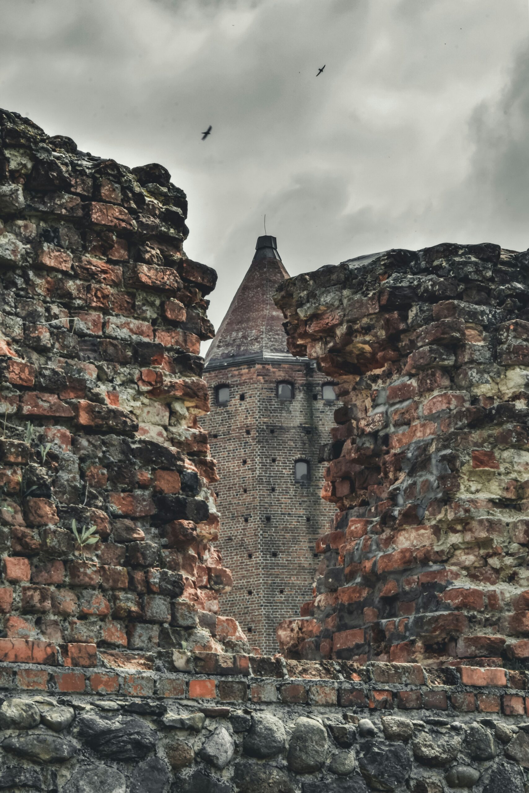 A medieval brick tower with a conical roof seen through the ruins of an old stone and brick wall, under an overcast sky with birds flying overhead.