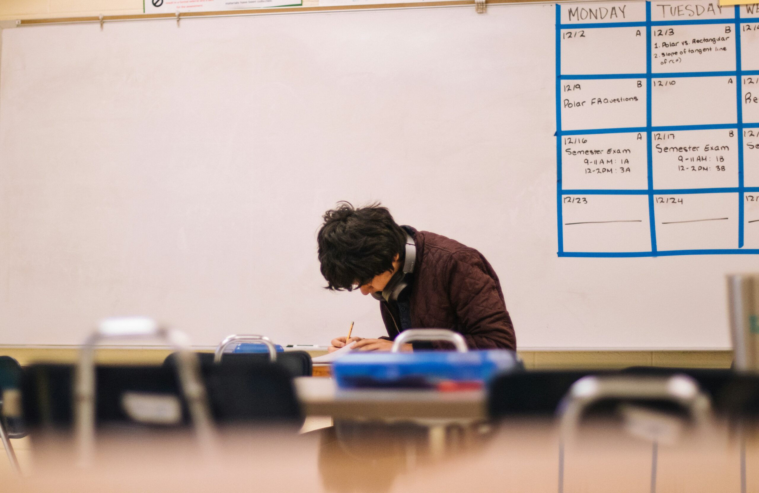 Student writing at a desk in an empty classroom with a large whiteboard calendar in the background.